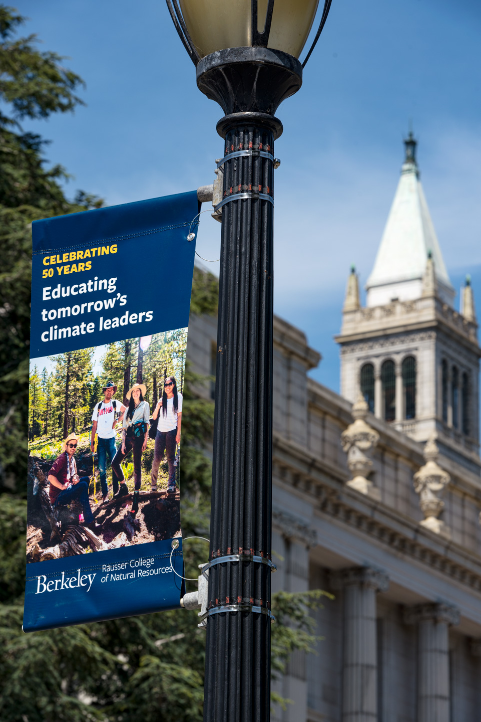 A photo of a banner featuring a group of smiling students is installed on a light post by outside. A large tower is visible in the background.