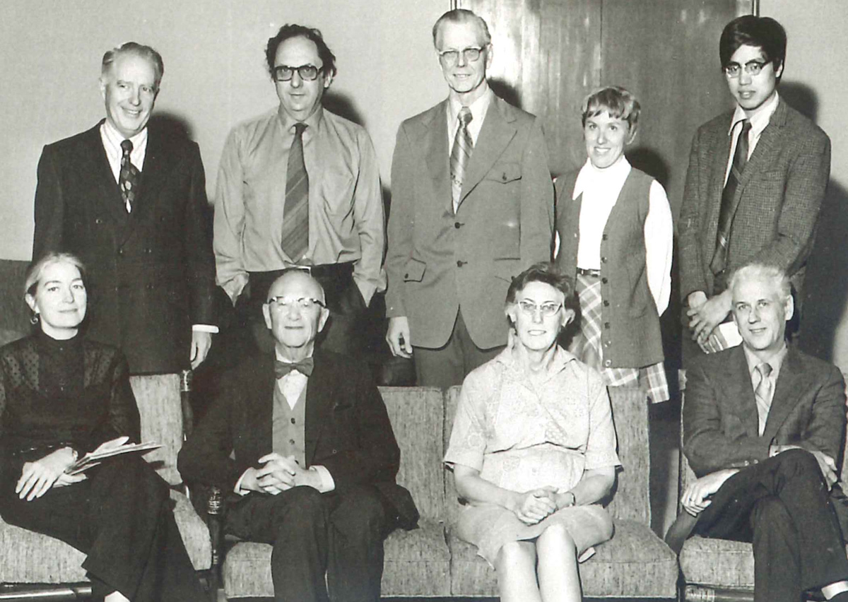A black and white photo of professors sitting and standing, smiling at the camera