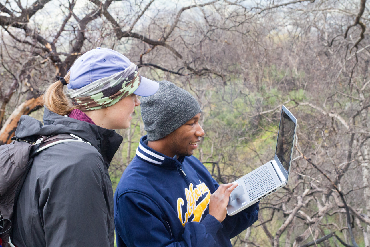 Jessie Moravek (left) and Kendall Calhoun (right). They are in an outdoor nature site, and Jessie is looking at Kendall who is holding a laptop.