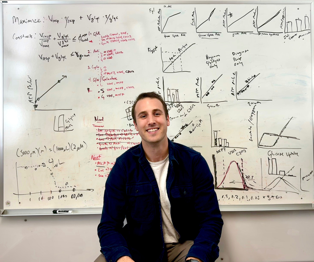 A photo of a smiling man in front of a whiteboard with equations behind him.