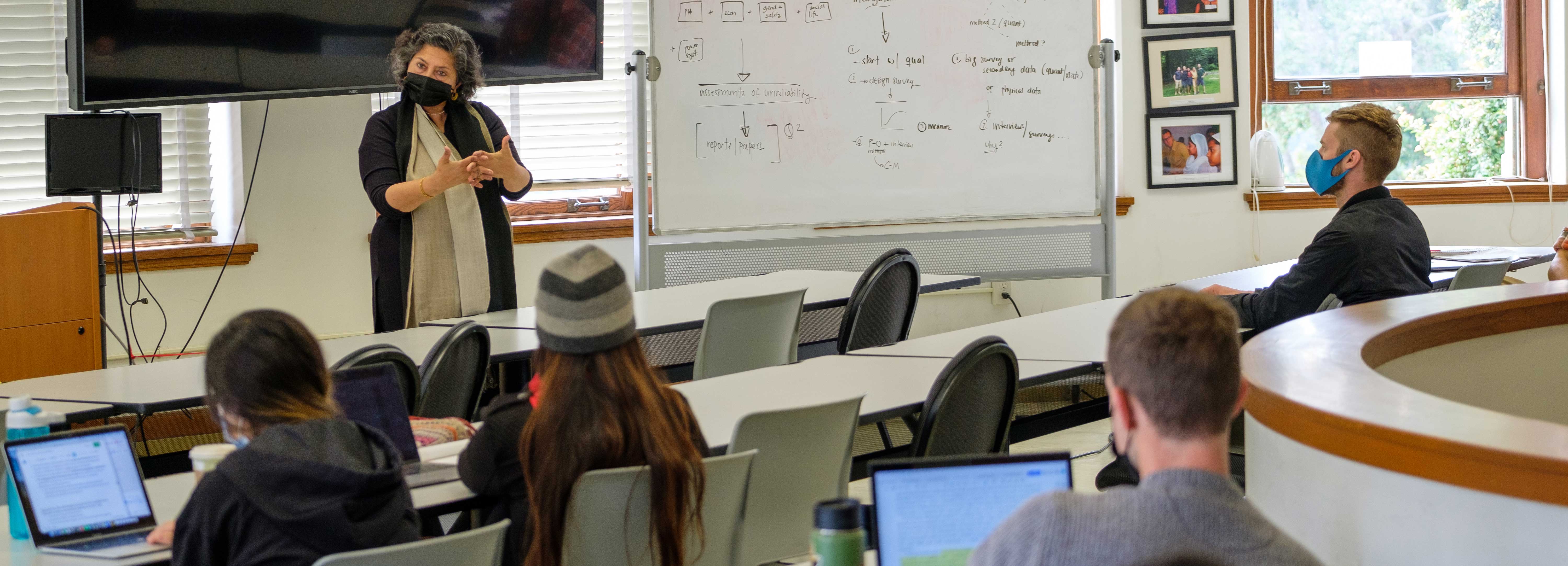 Isha Ray next to a whiteboard, speaking to a class