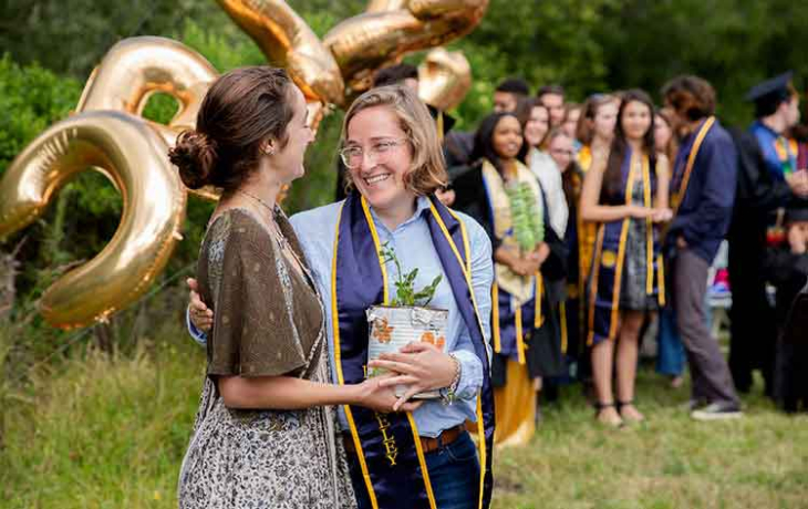 Two young graduates stand holding a potted plant with other graduating seniors behind them