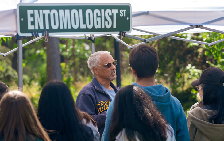 Vernard Lewis with a group of students facing him. They are under a tarp with a sign that says "Entomologist St" atop the tarp. Behind them is a forest.