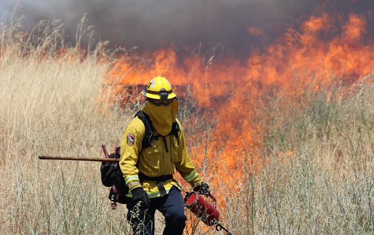 A person in yellow protective gear lights a fire in tall grass.