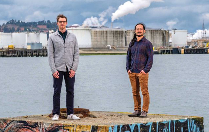 Nicholas Depsky and Seigi Karasaki stand in front of a factory near the coast
